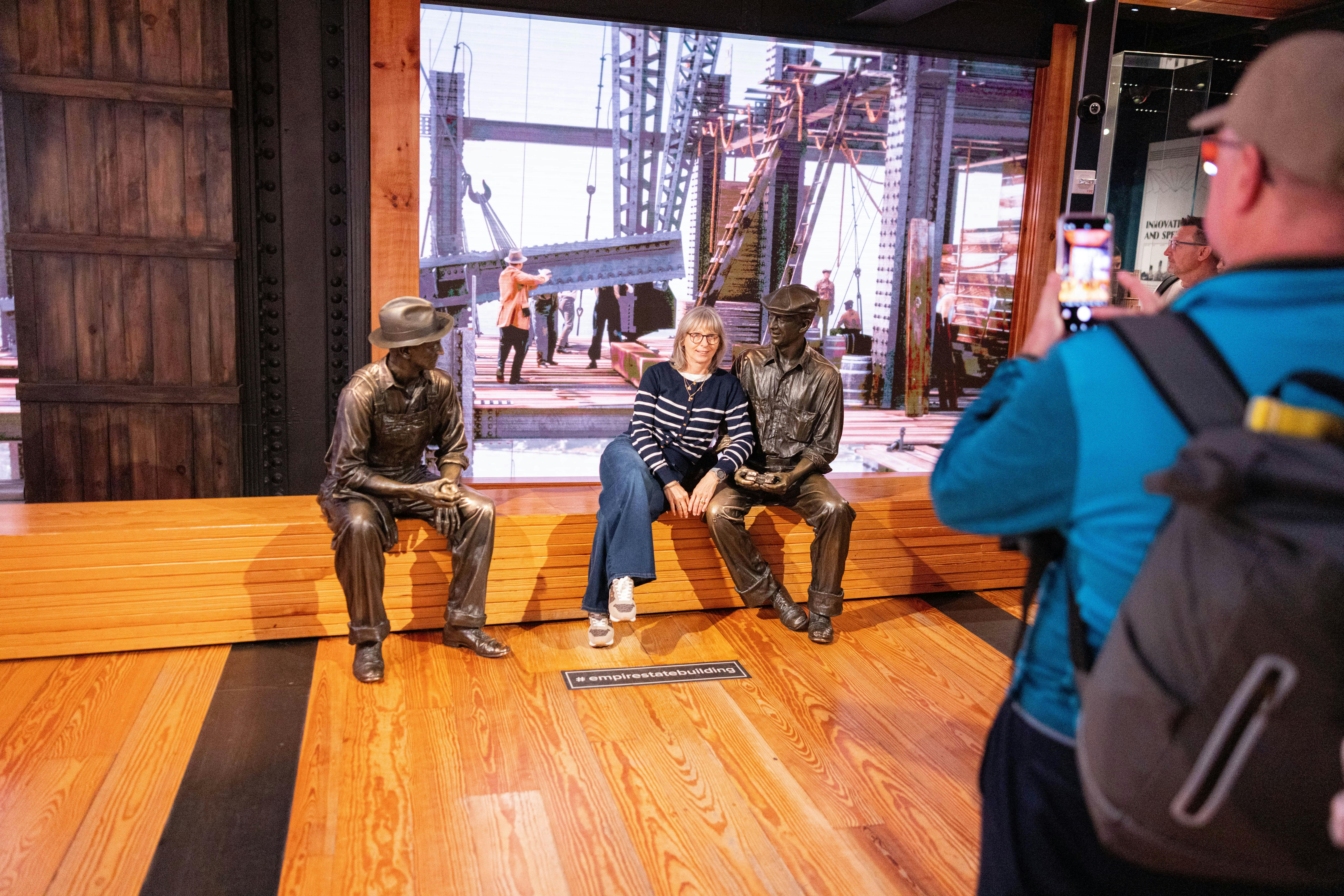A woman sitting between two bronze statues of workers on a wooden bench, with a construction scene displayed behind.