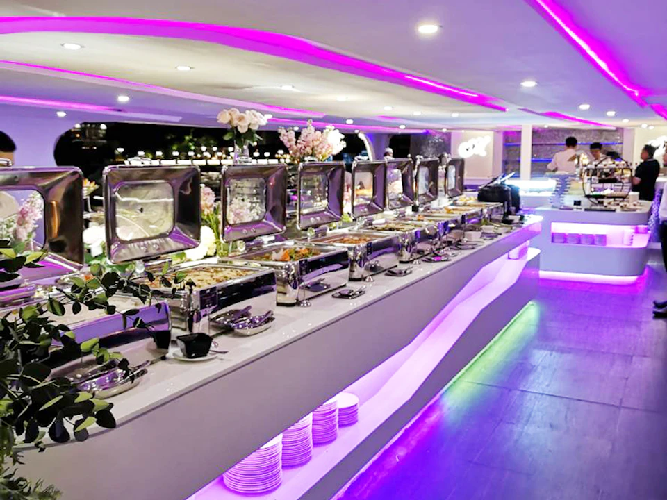 Long buffet table with multiple food trays under purple lighting, decorated with flowers and stacked plates.