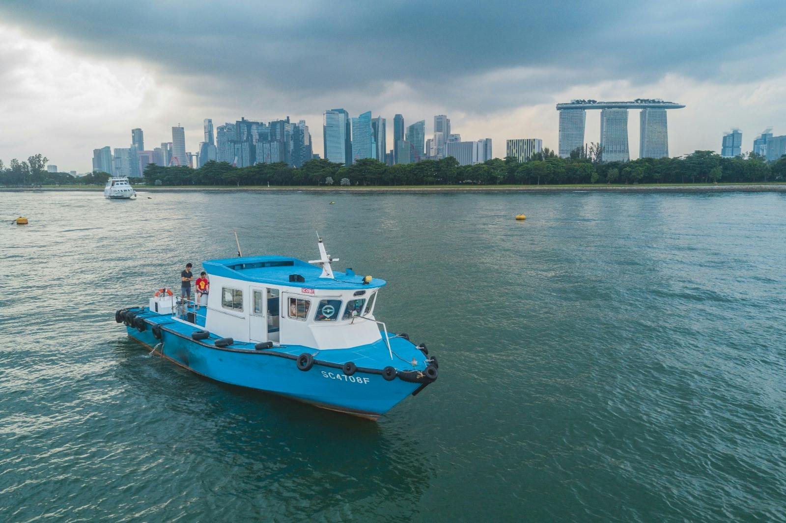 A blue and white boat on water with a city skyline and the Marina Bay Sands hotel in the background under a cloudy sky.