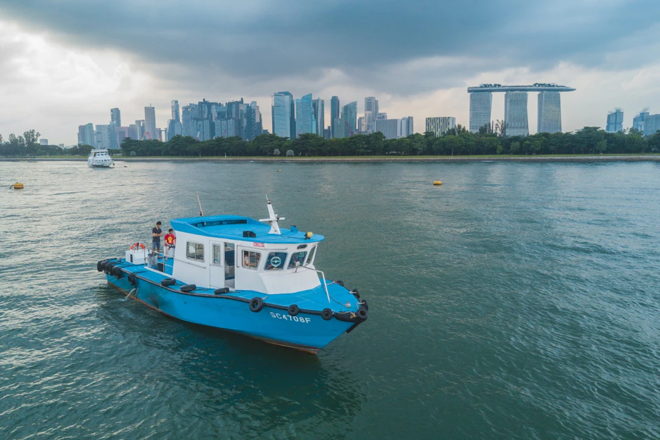 A blue and white boat on water with a city skyline and the Marina Bay Sands hotel in the background under a cloudy sky.