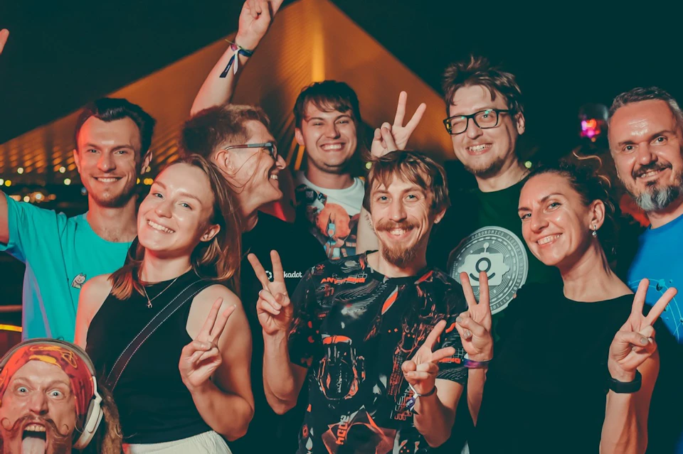 A group of people smiling and making peace signs, with a night scene and a lit triangular structure in the background.