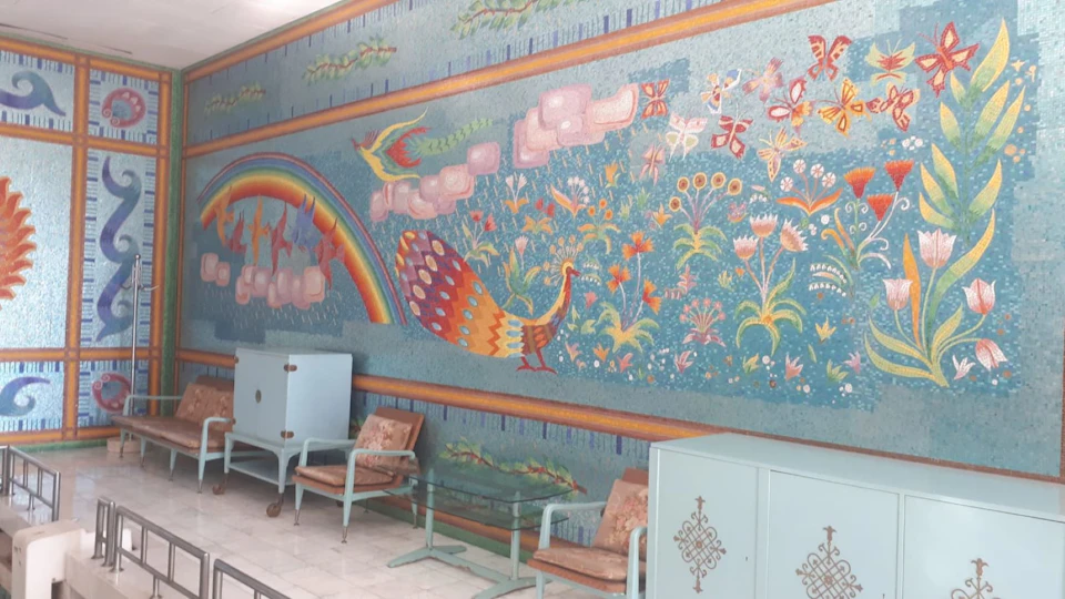 Colorful mural featuring peacocks, flowers, and a rainbow on a blue wall, with light blue cabinets and chairs arranged below.
