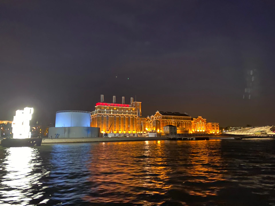 Illuminated waterfront building and cylindrical structure reflecting light in the water at night under a dark sky.