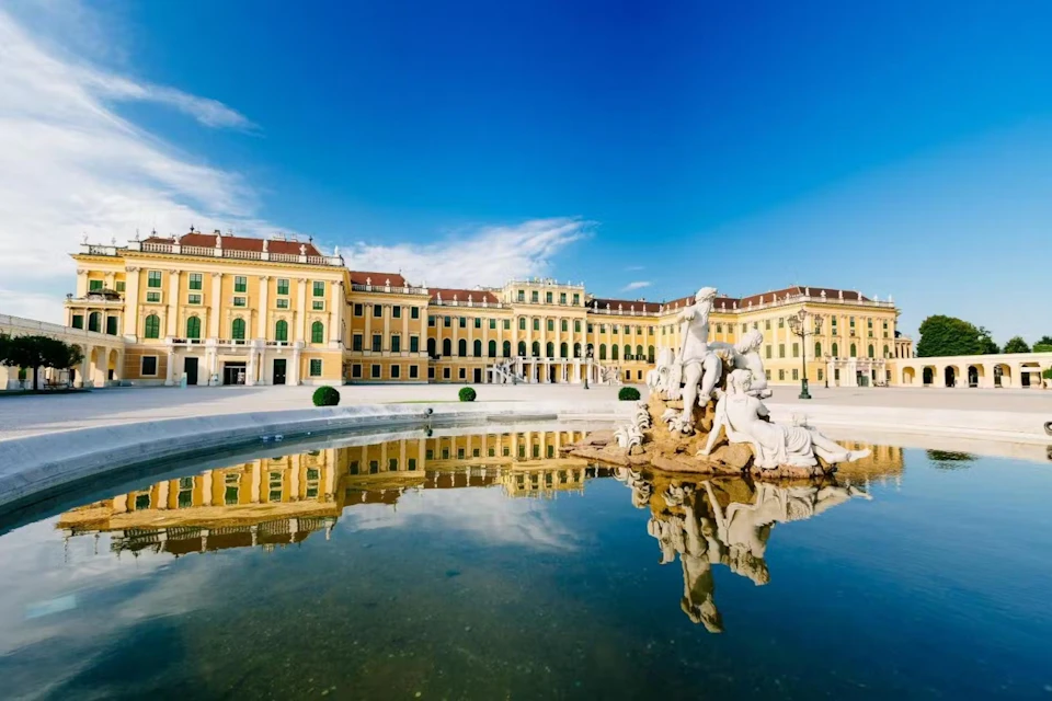 Large yellow palace with a statue fountain in the foreground, reflecting in the water, under a clear blue sky.