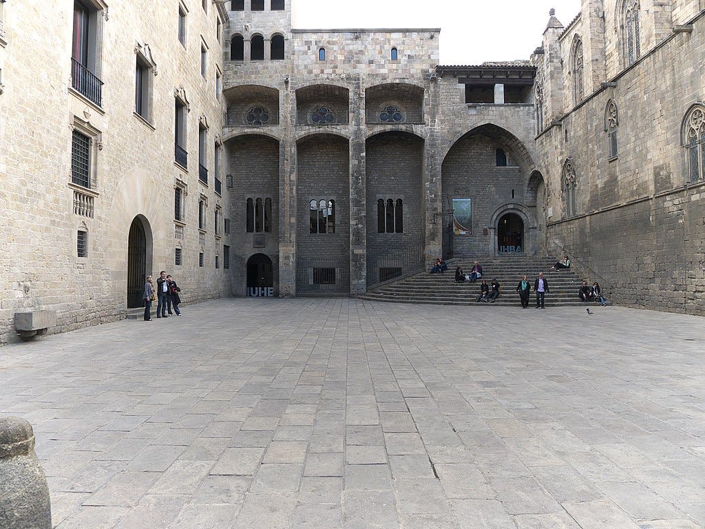 A large stone courtyard surrounded by historic buildings with arched windows and doors; a few people are sitting and walking.