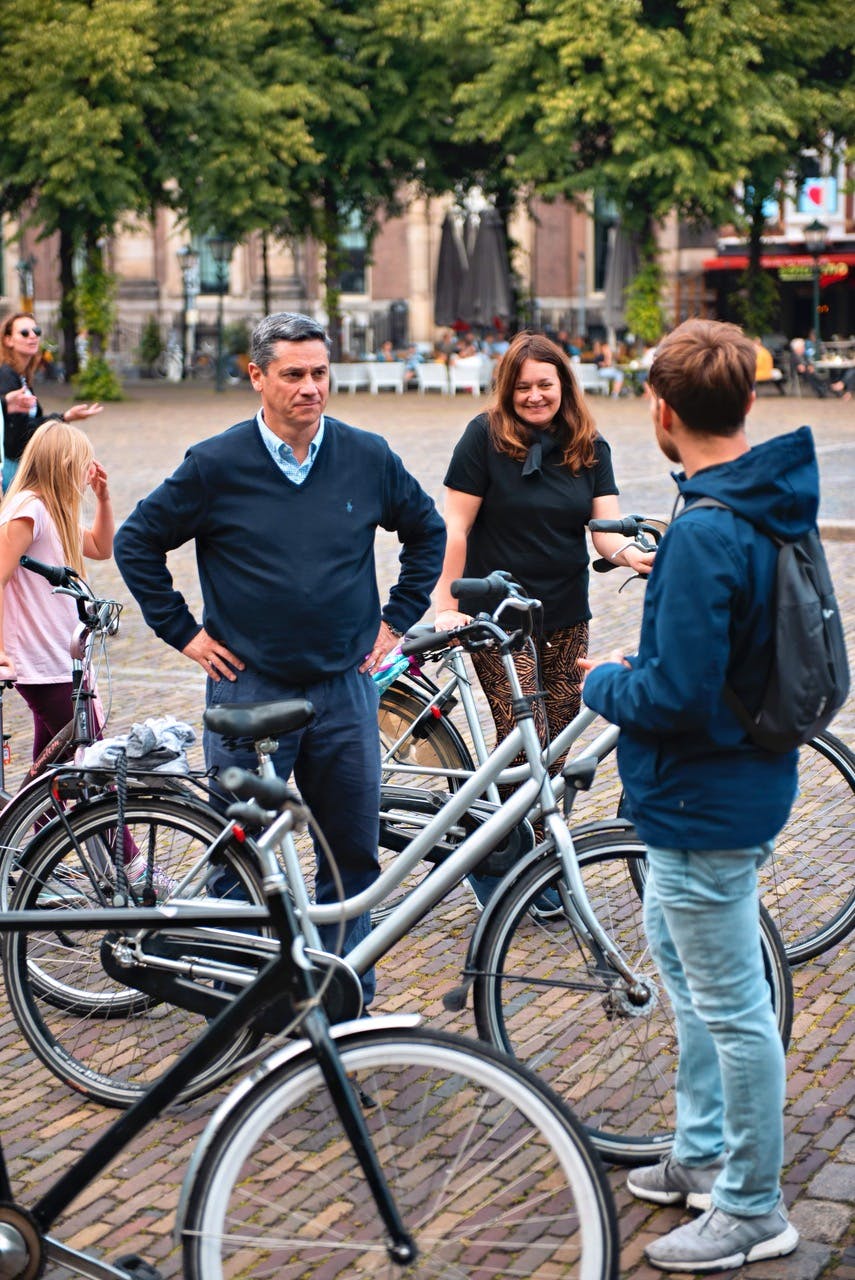 Un gruppo di persone si trova con le biciclette su una strada acciottolata, con alberi ed edifici sullo sfondo. Due adulti stanno conversando.