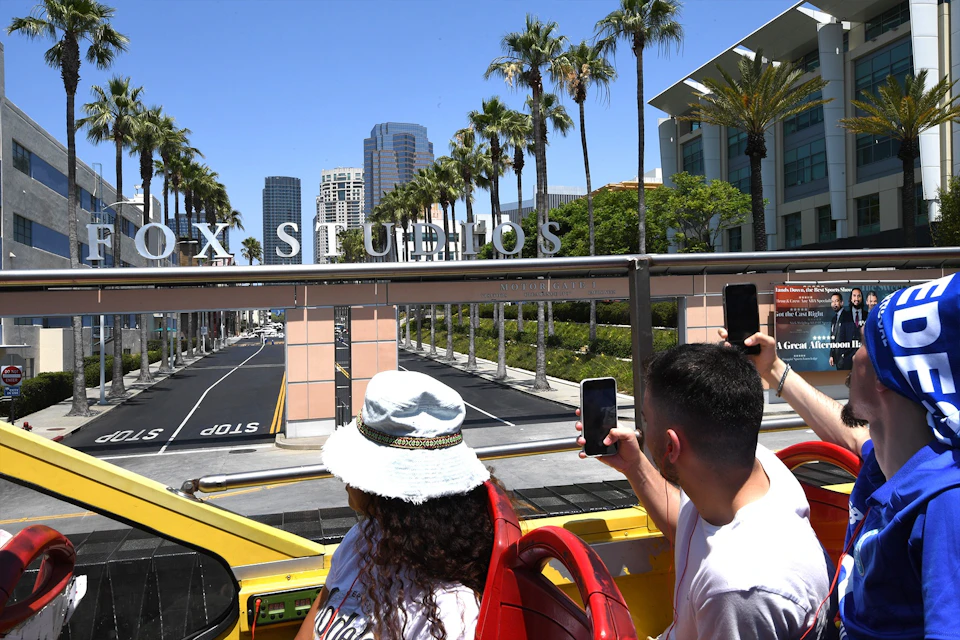 Tourists on an open-top bus taking photos of the Fox Studios sign with palm trees and buildings in the background.