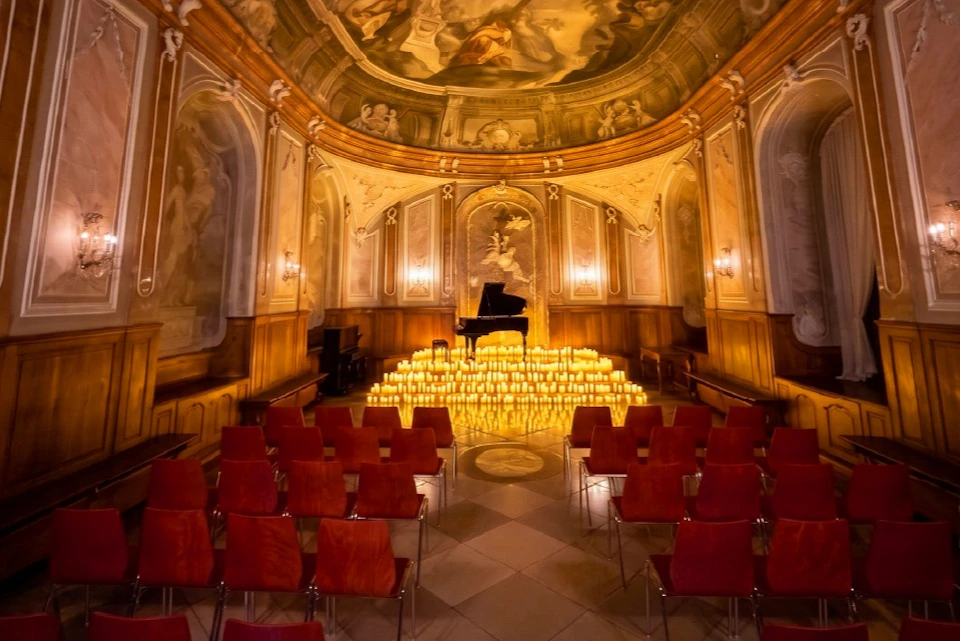 A grand room with a piano on a stage surrounded by numerous lit candles, viewed from rows of empty red chairs.
