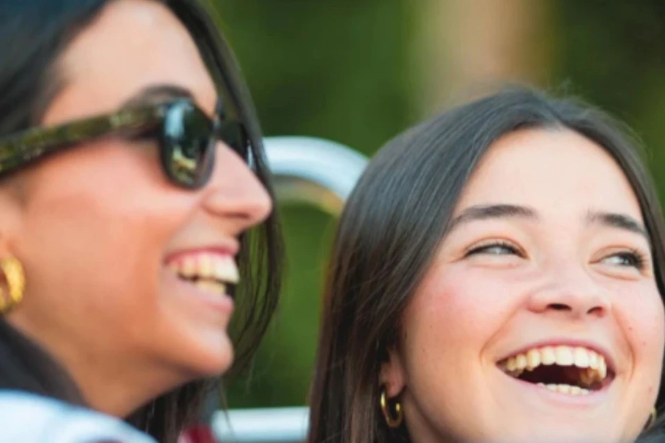 Two women smiling and laughing outdoors, with blurred greenery in the background.