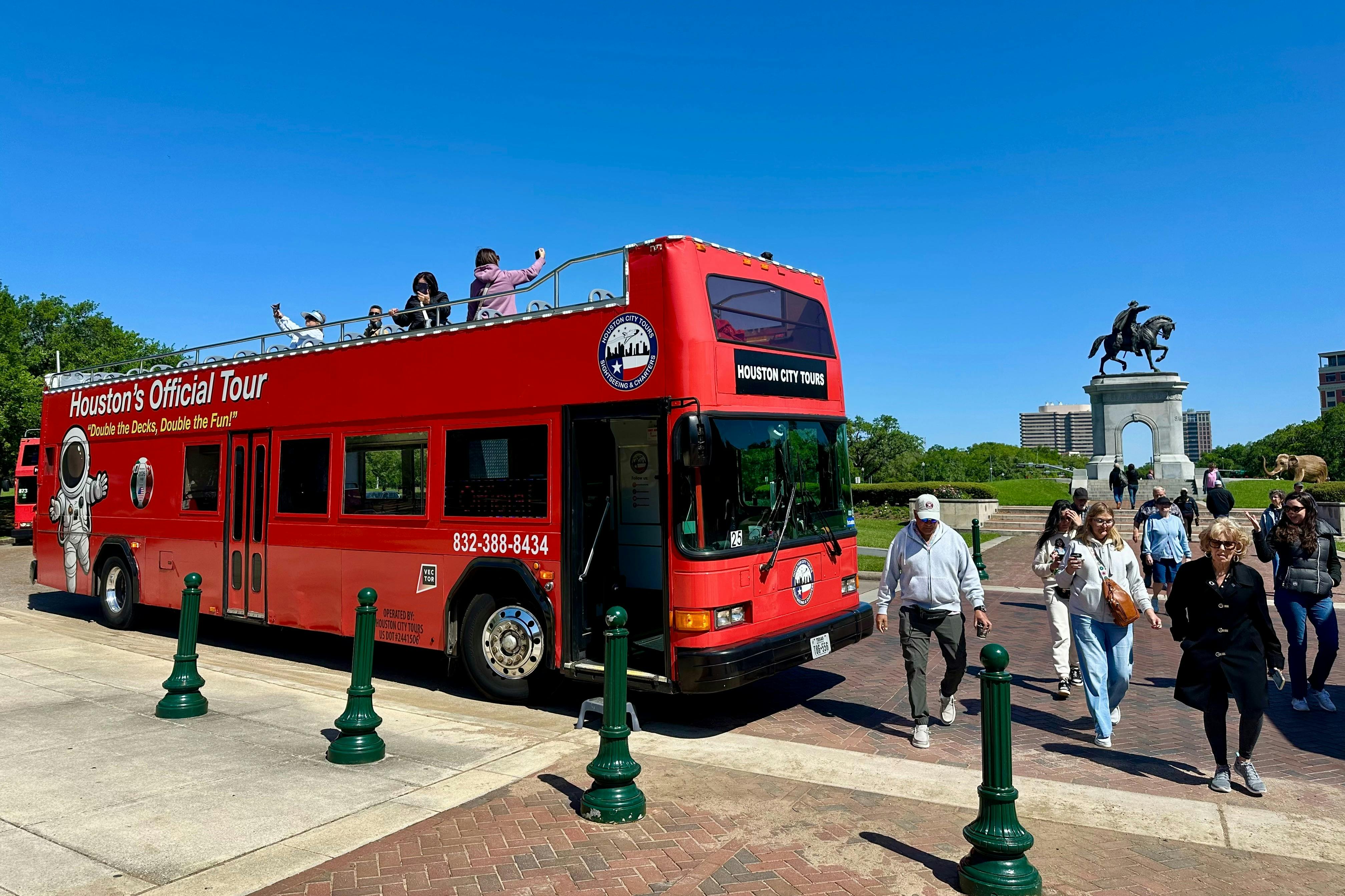 A red double-decker tour bus parked near a statue of a rider on horseback, with people walking around under a clear blue sky.
