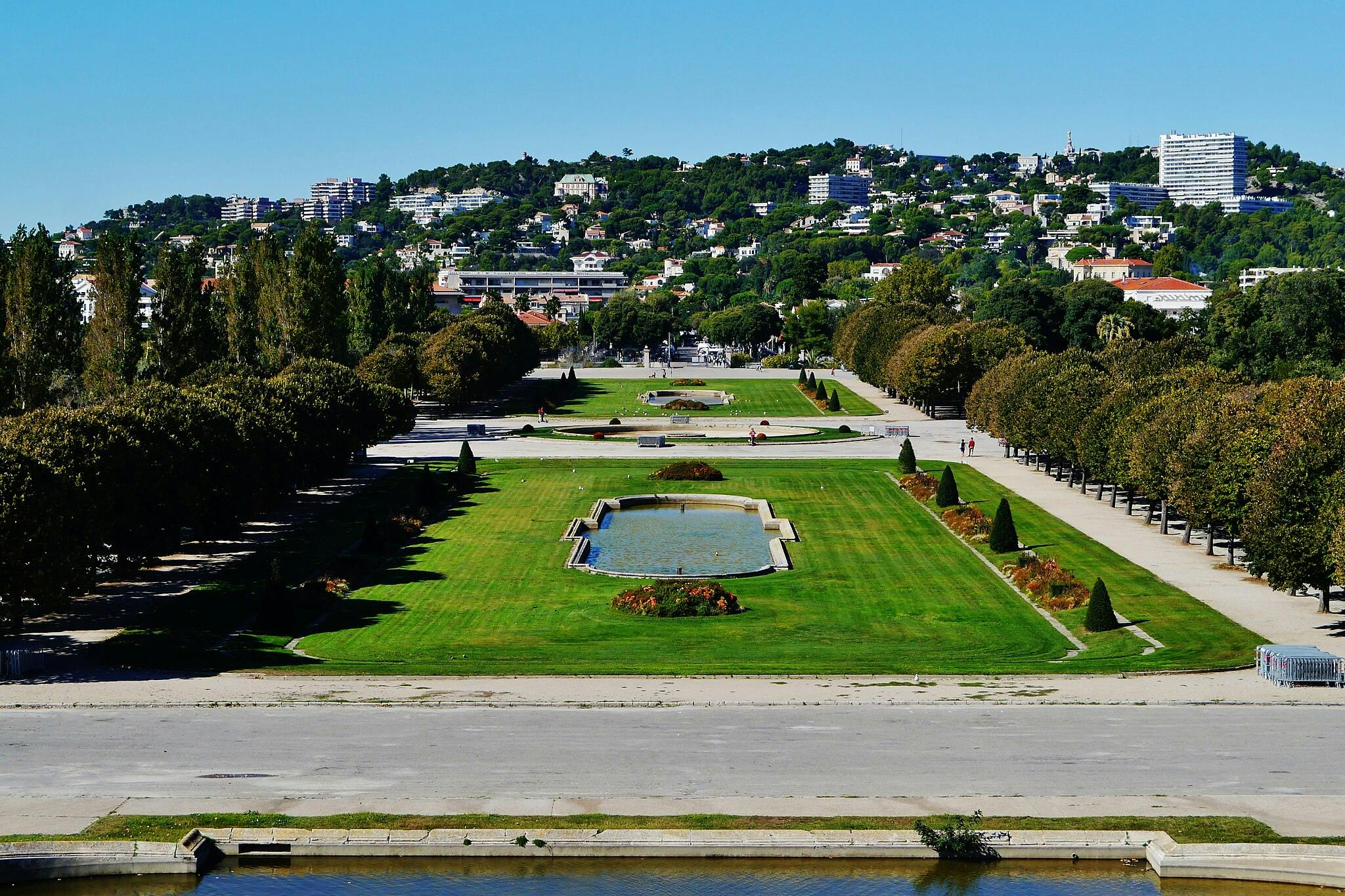 잘 다듬어진 잔디밭, 직사각형 연못, 통로, 나무, 언덕 마을의 먼 풍경이 있는 조경 공원입니다.