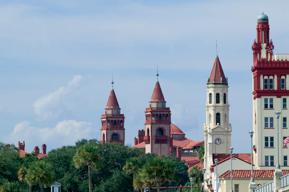 Historic buildings with red-tiled roofs and spires, partially obscured by trees, set against a blue sky with light clouds.