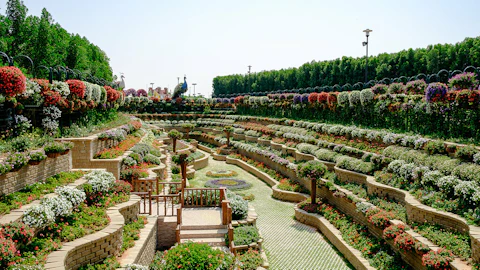 Terraced garden with cascading flower beds, colorful flowers, and neatly trimmed bushes. Pathways and a wooden gazebo are visible.