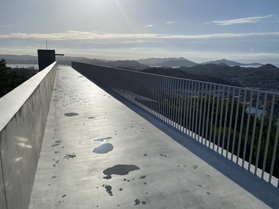 A concrete walkway with puddles, bordered by a metal railing, overlooking a mountainous landscape under a bright sky.