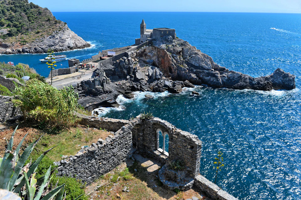 Ancient stone structures on a rocky coastline with a church on a cliff, overlooking the blue ocean under a clear sky.