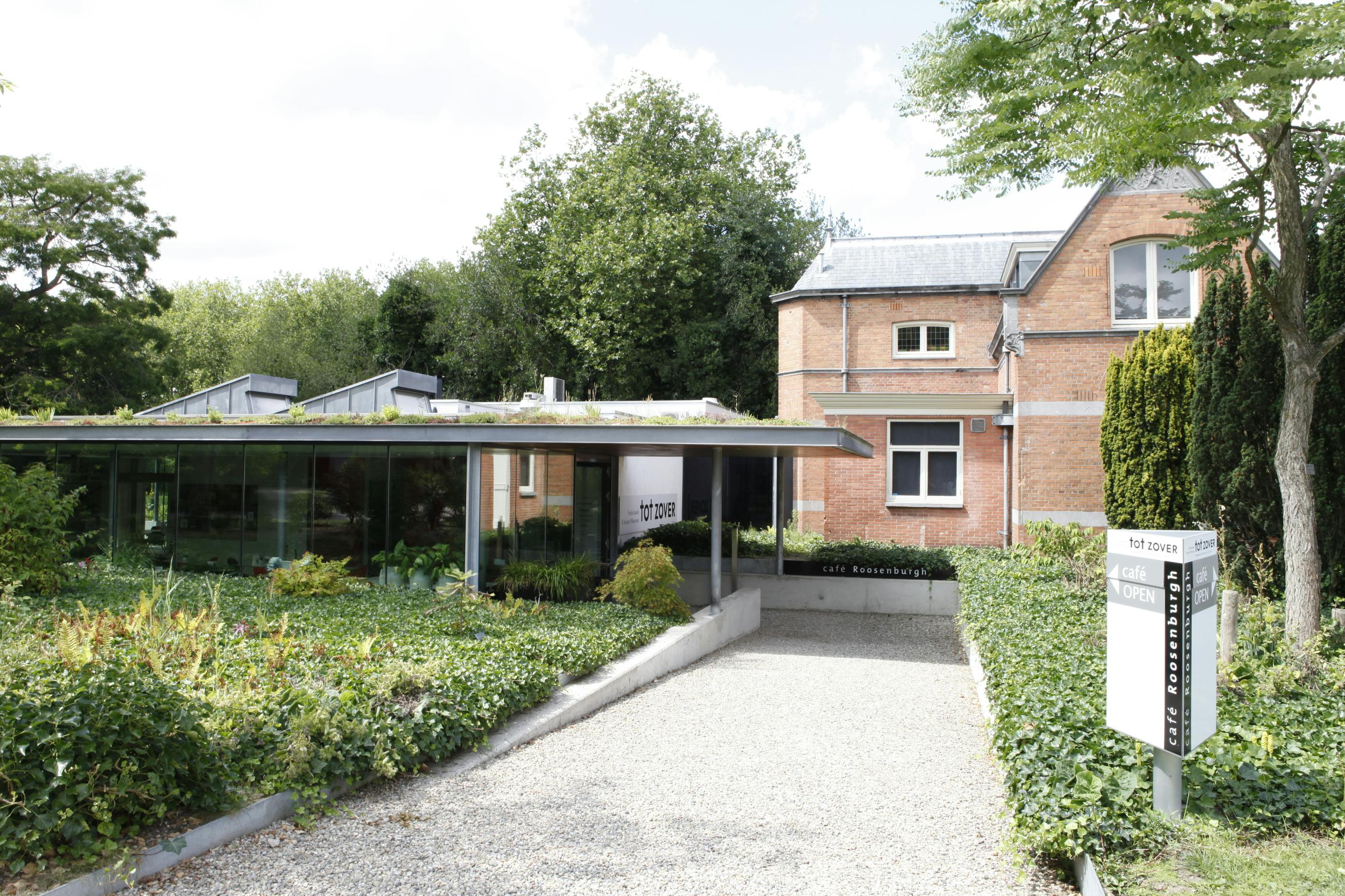 Un modern edifici amb parets de vidre al costat d'una casa de maó, envoltat de vegetació amb un rètol que diu "cafè Roosenburgh" i "tot zover".