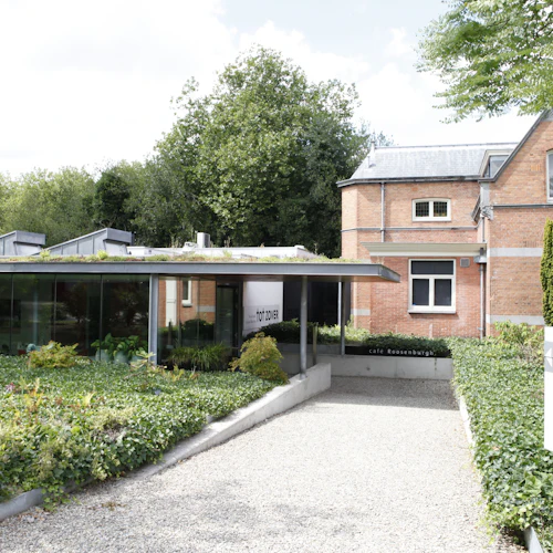En modern byggnad med glasväggar bredvid ett tegelhus, omgiven av grönska med en skylt där det står ”cafe Roosenburgh” och ”tot zover”.