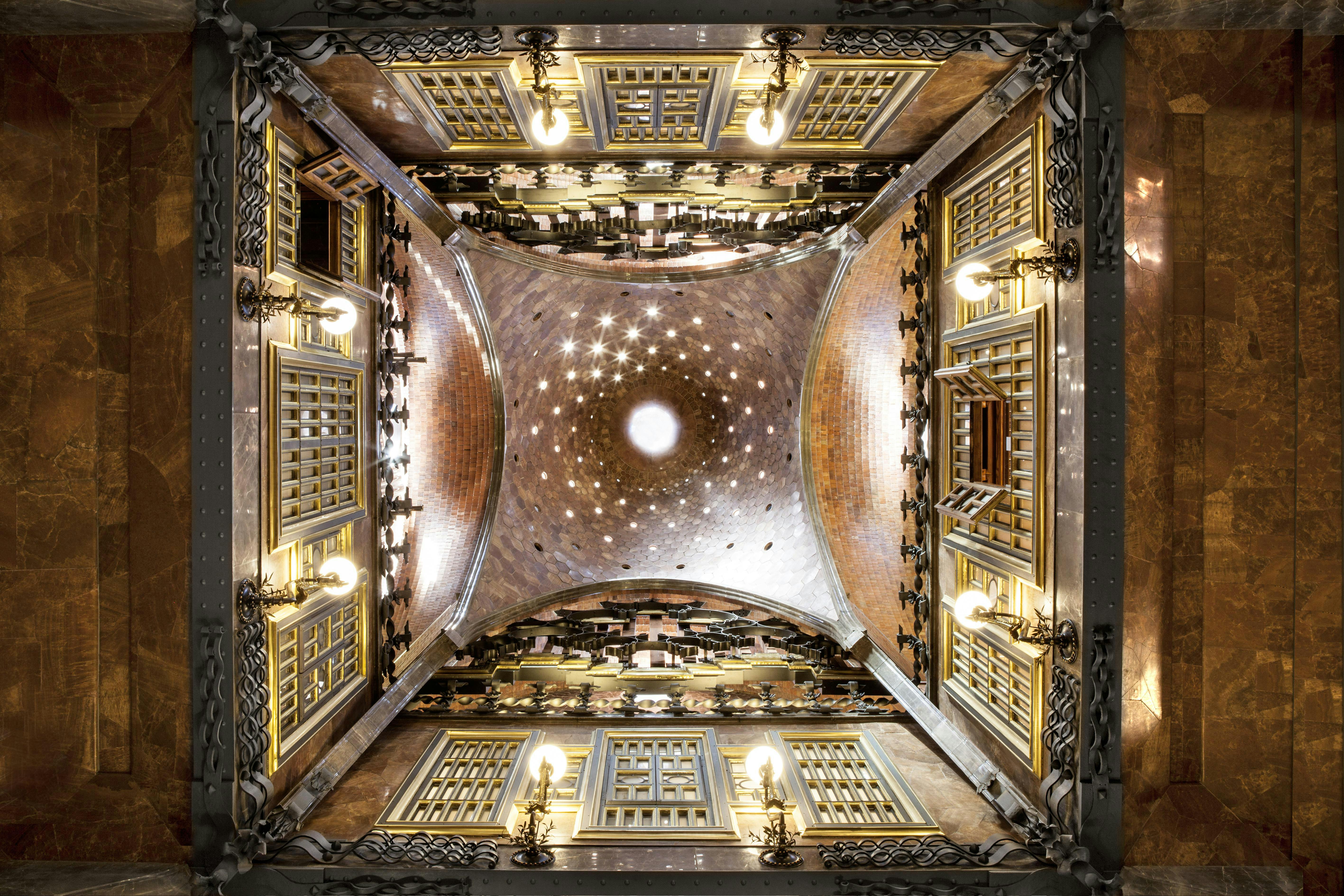 A symmetrical shot of an ornate, vaulted ceiling with decorative lights, windows, and intricate architectural detailing.