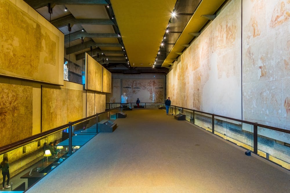 博物馆中有宽敞的走廊，有大型壁画和聚光灯，有两个人走路，一个人坐在远处。