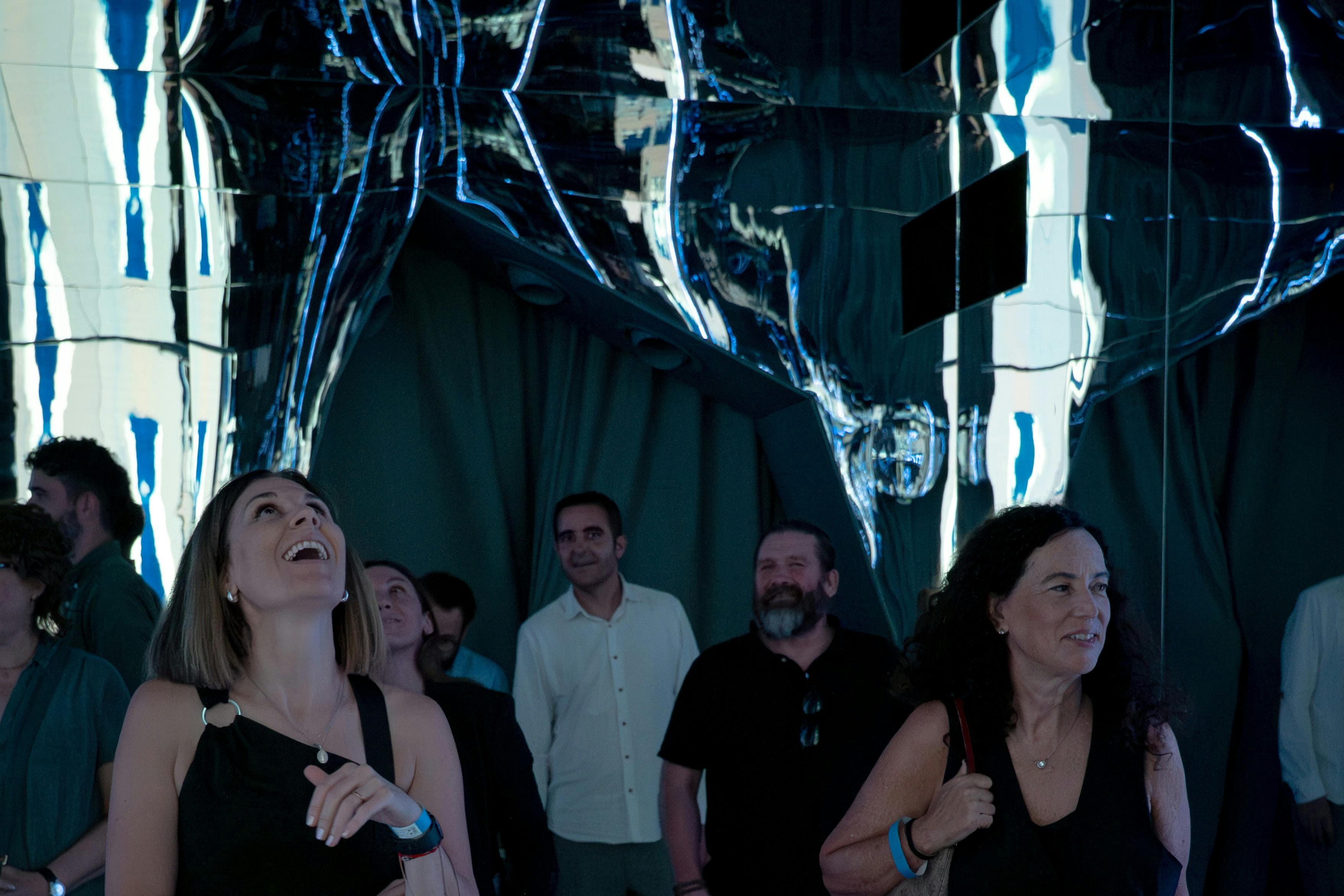 Skupina lidí stojí pod reflexním, zkresleným kovovým uměním ve slabě osvětlené místnosti se záclonami.