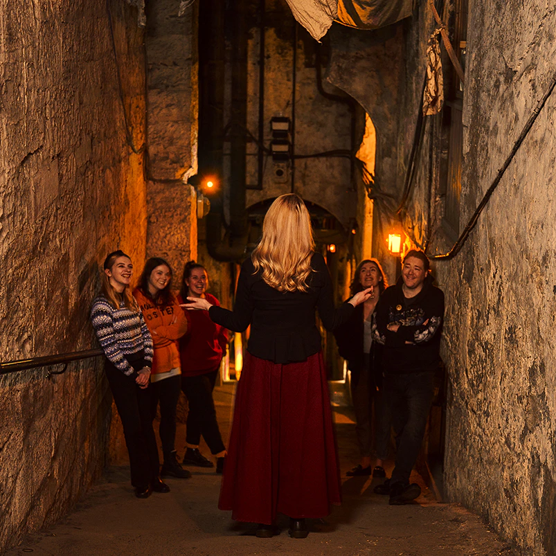 A woman in a red skirt leads a small tour group in a dimly lit, narrow stone alley. The group listens attentively.