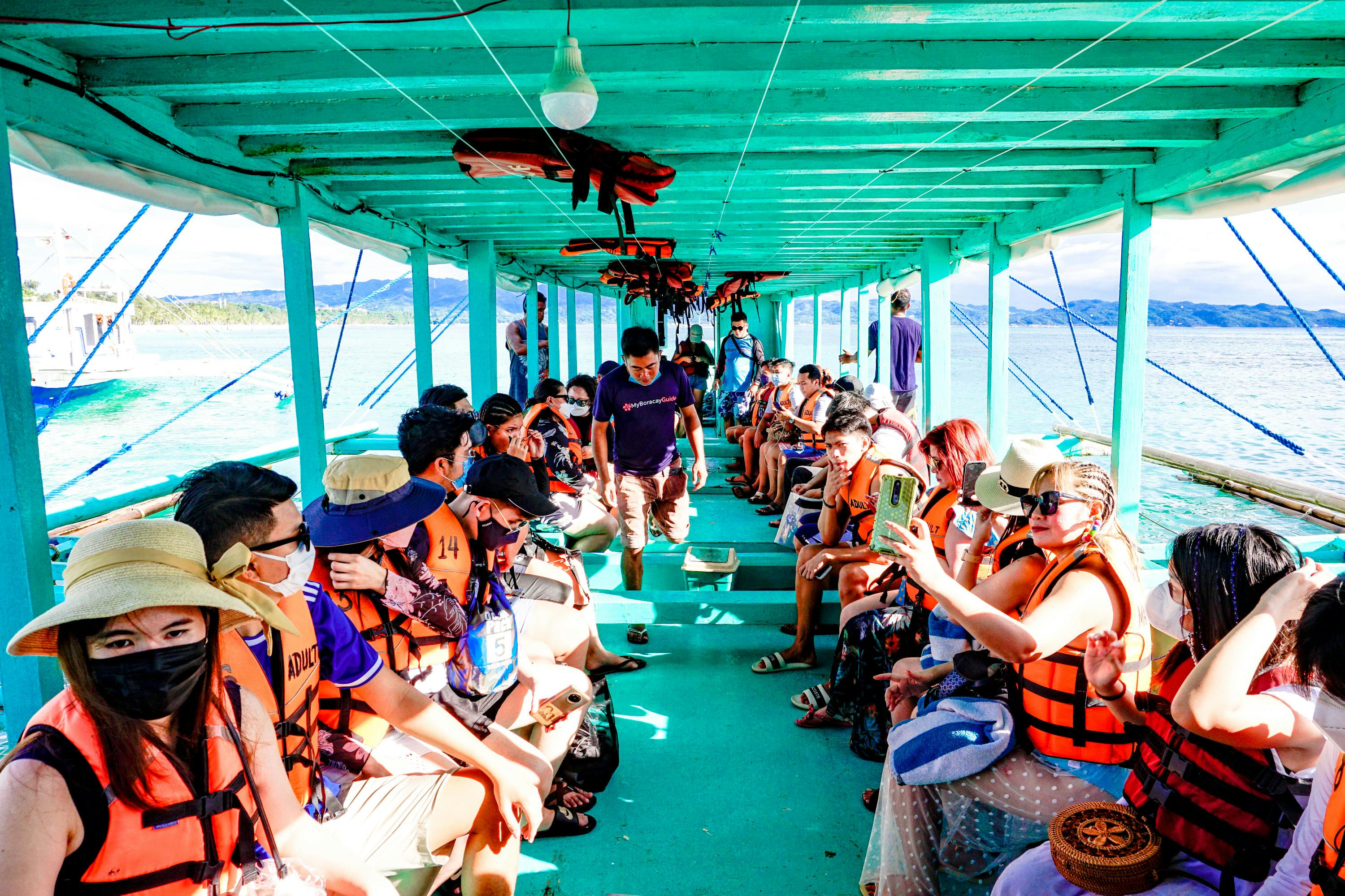 A group of people wearing life jackets seated inside a turquoise-painted boat on a sunny day, with water visible outside.