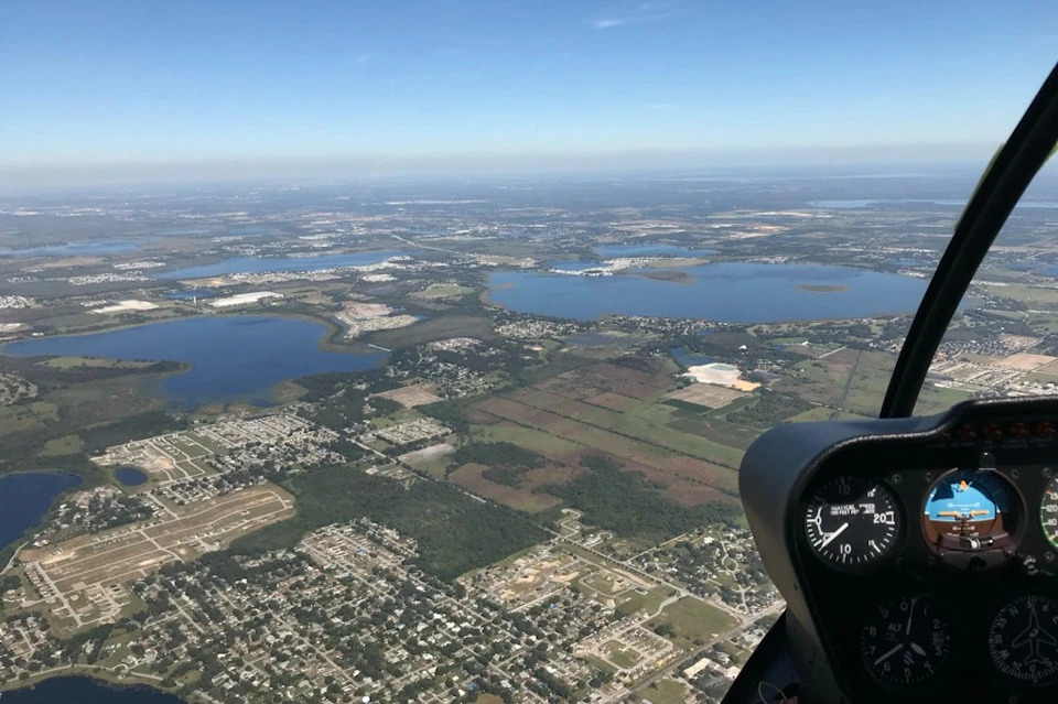 Aerial view of a landscape with lakes, fields, and urban areas. Part of an aircraft instrument panel is visible in the foreground.