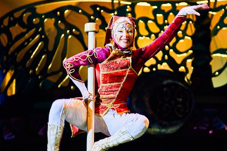 Una intèrpret vestida amb un vestit de colors posa a l'escenari amb un pal, contra un teló de fons decoratiu.