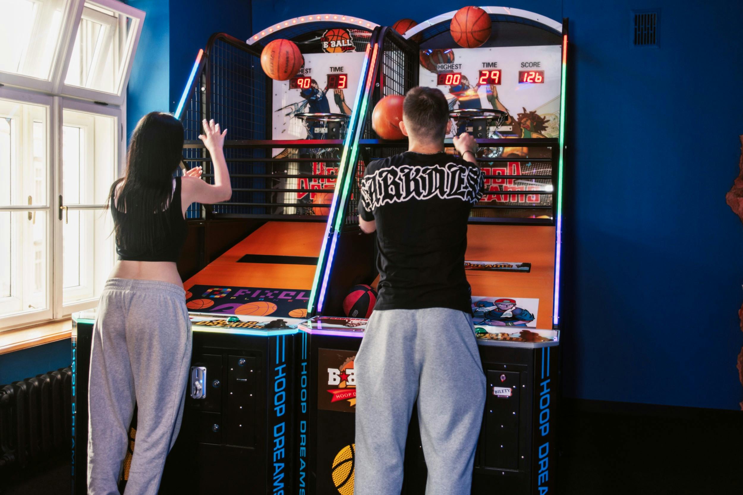 Two people playing an arcade basketball game, aiming to shoot basketballs into hoops under colorful lighting.