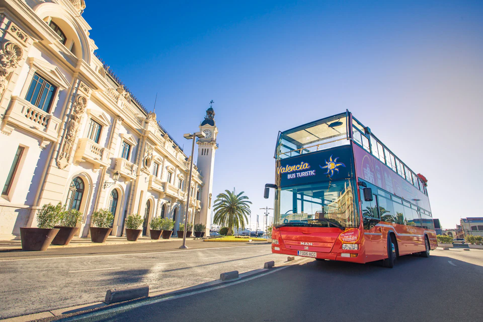 A red double-decker tour bus labeled "Valencia Bus Turistic" is parked beside a historic white building and palm trees on a sunny day.