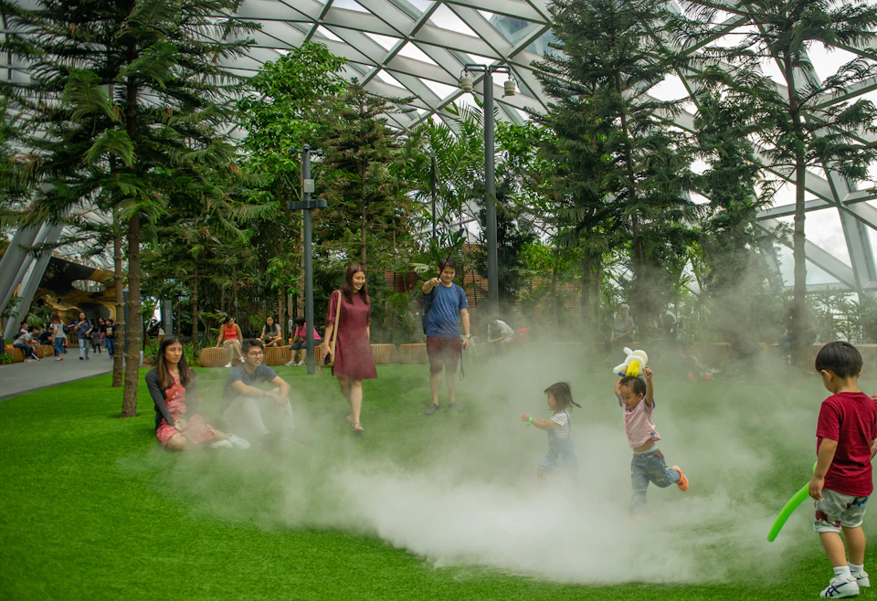 People relaxing and walking on grass in a large indoor garden with trees and mist under a geometric glass roof.