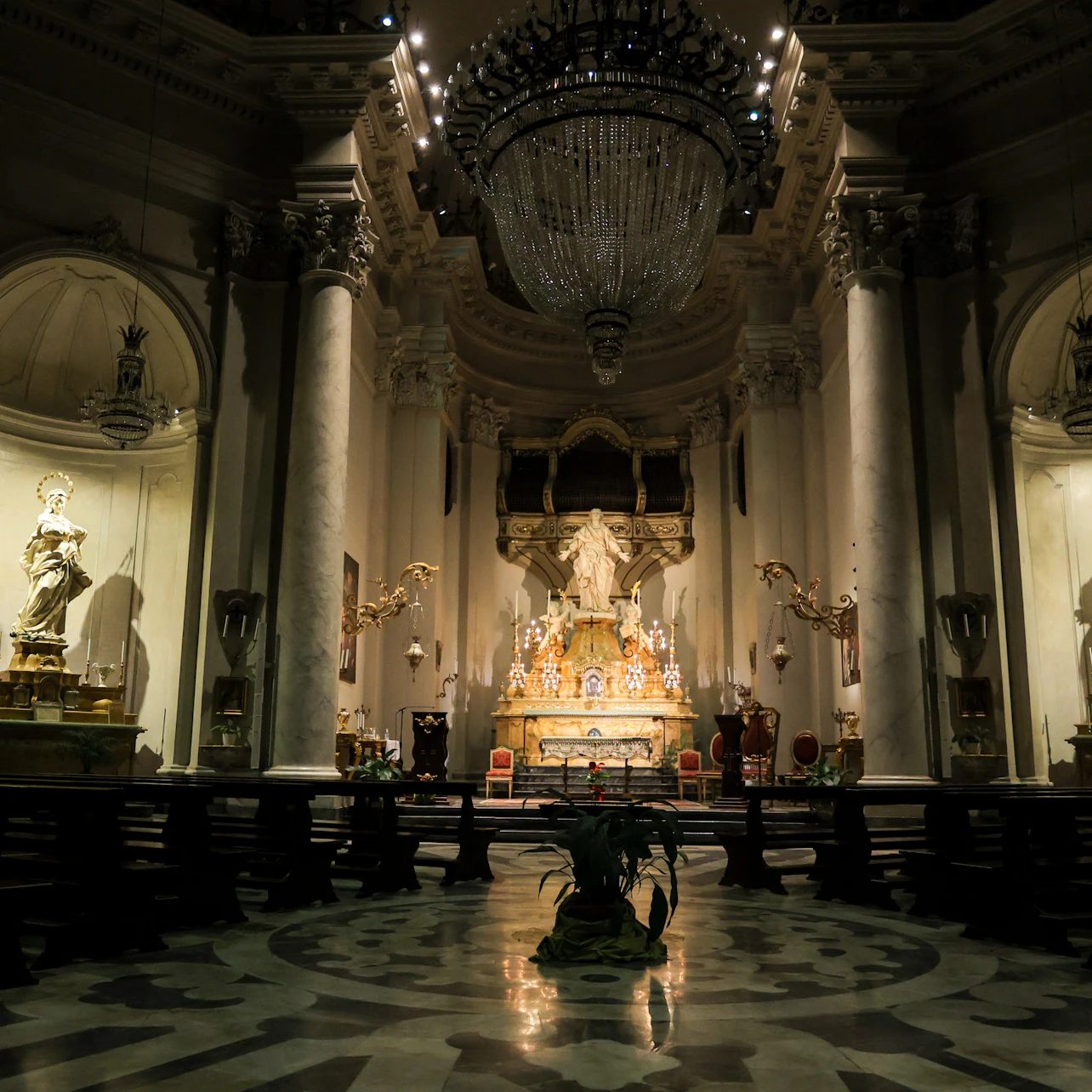 A dimly lit, ornate church interior with a large chandelier, statues, an altar, and intricate architectural details.