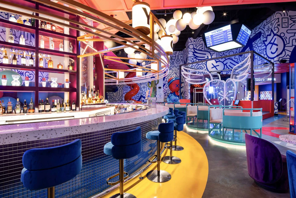 A vibrant modern bar with blue stools, colorful decor, and artistic murals. Seating areas and a digital menu are visible.