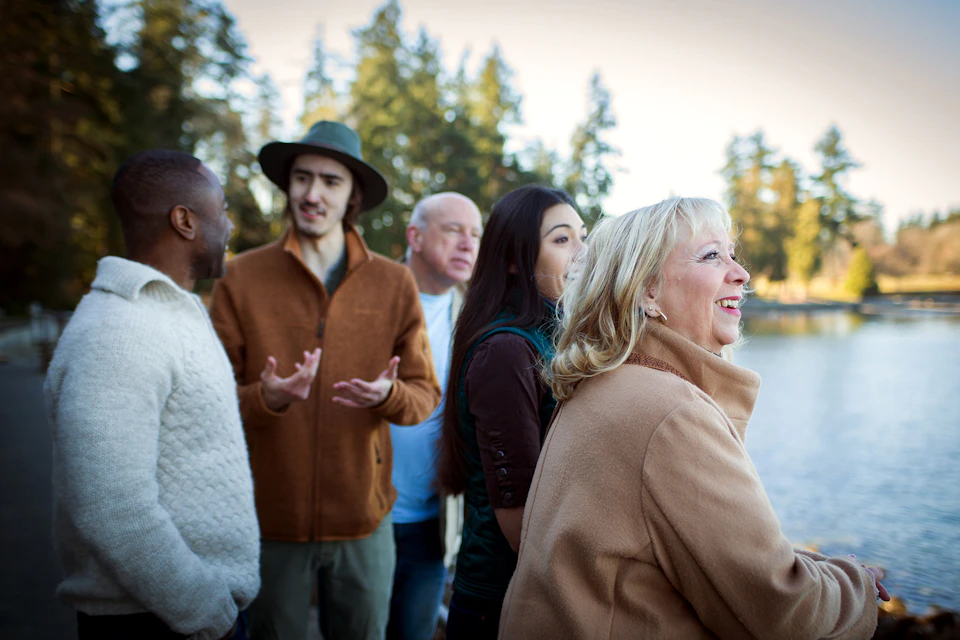 A group of people outdoors in casual attire, talking and smiling, with trees in the background.