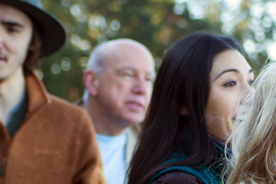 A group of people outdoors in casual attire, talking and smiling, with trees in the background.