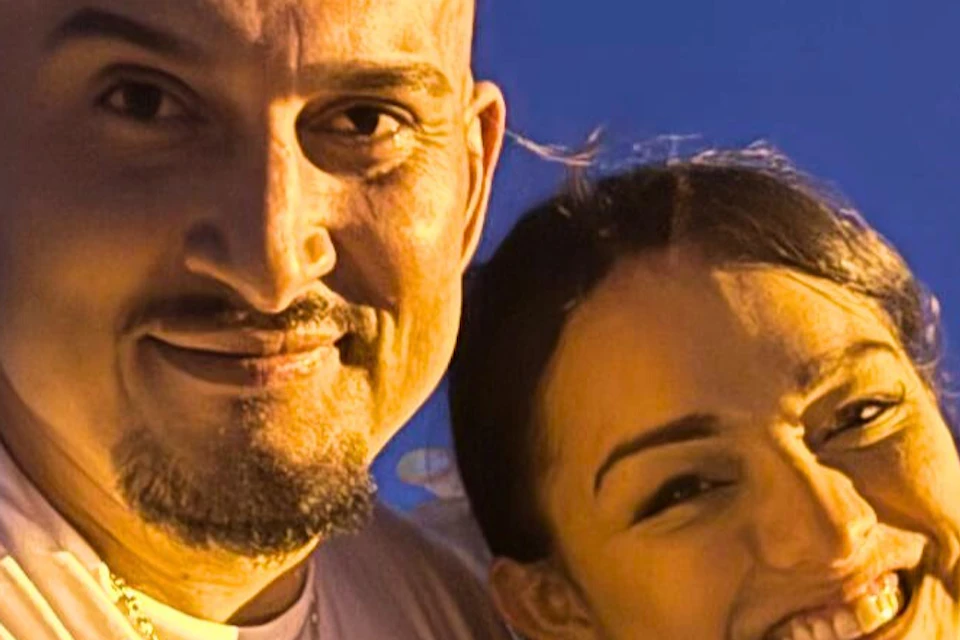A man and woman are smiling and pointing at the camera. The woman is holding a red heart-shaped balloon.