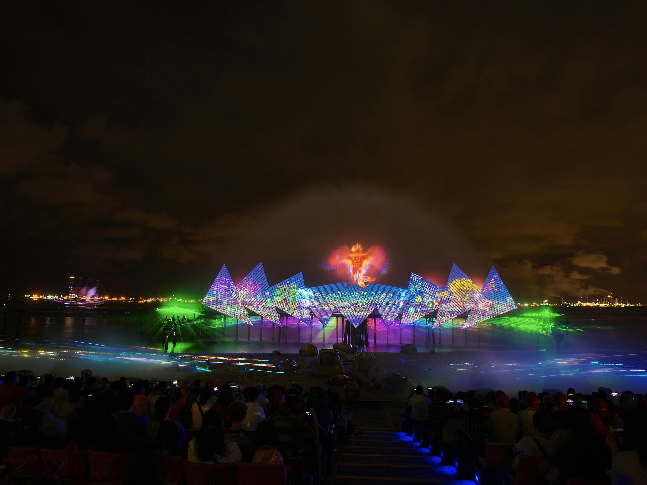 Noční venkovní show se zářivými světly a projekcemi na vodních a trojúhelníkových obrazovkách, kterou uvidí sedící diváci.