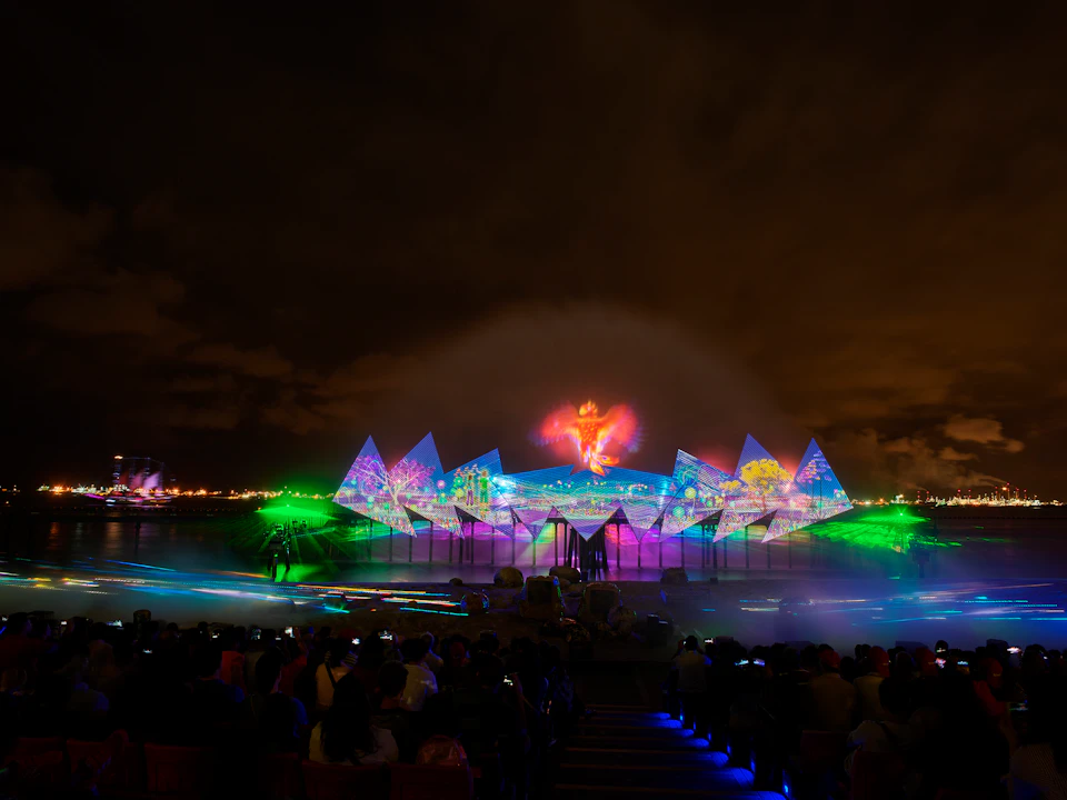 Nighttime outdoor show with vibrant lights and projections on water and triangular screens, viewed by a seated audience.