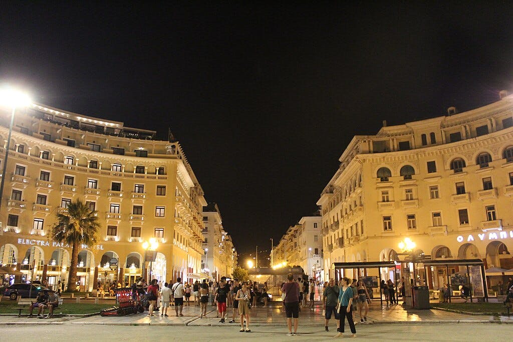 People walking in a well-lit plaza at night, bordered by grand buildings, including the Electra Palace Hotel.