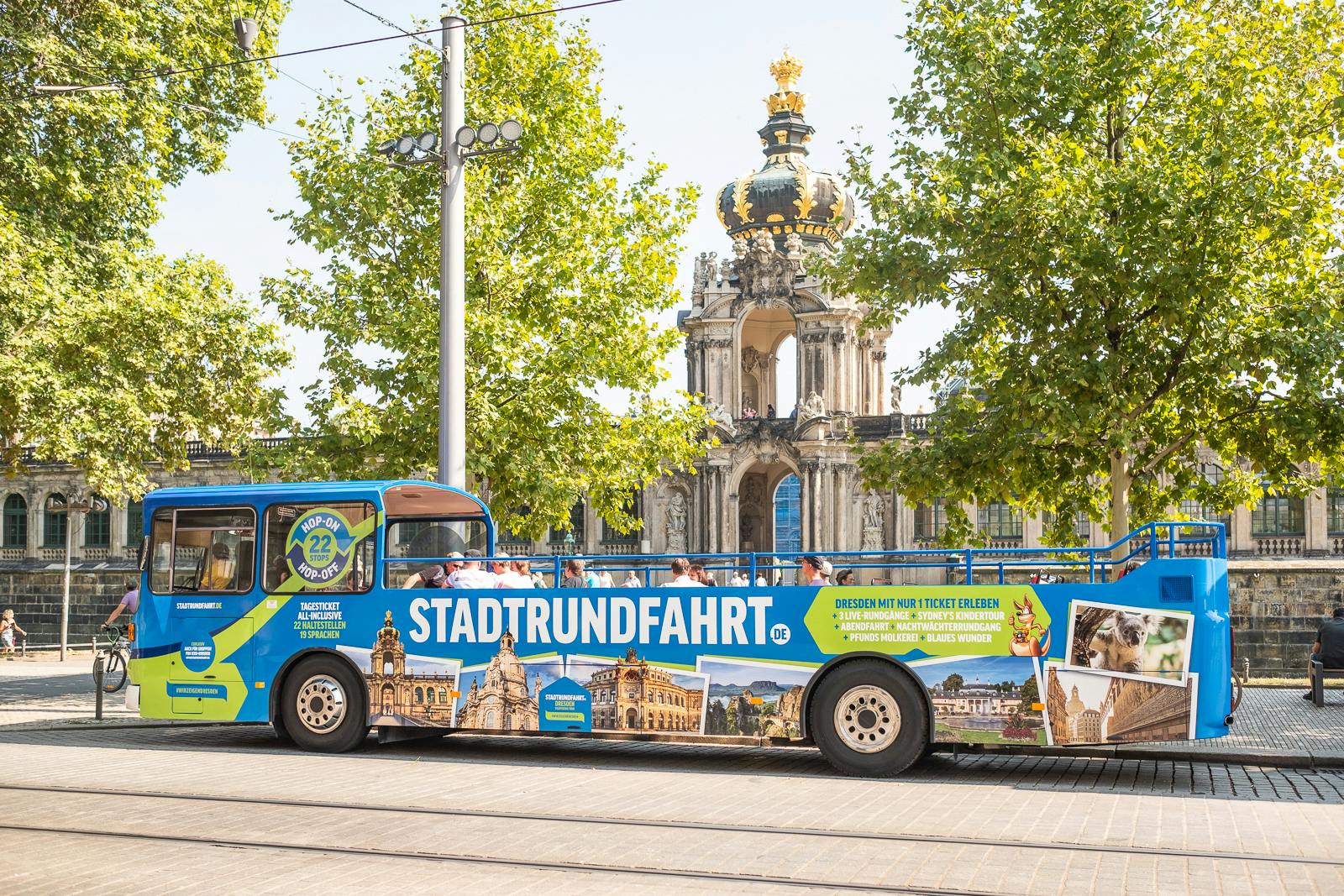 Blue tour bus with "STADTRUNDFAHRT" signage parked in front of a historic building with a gold dome, surrounded by trees.