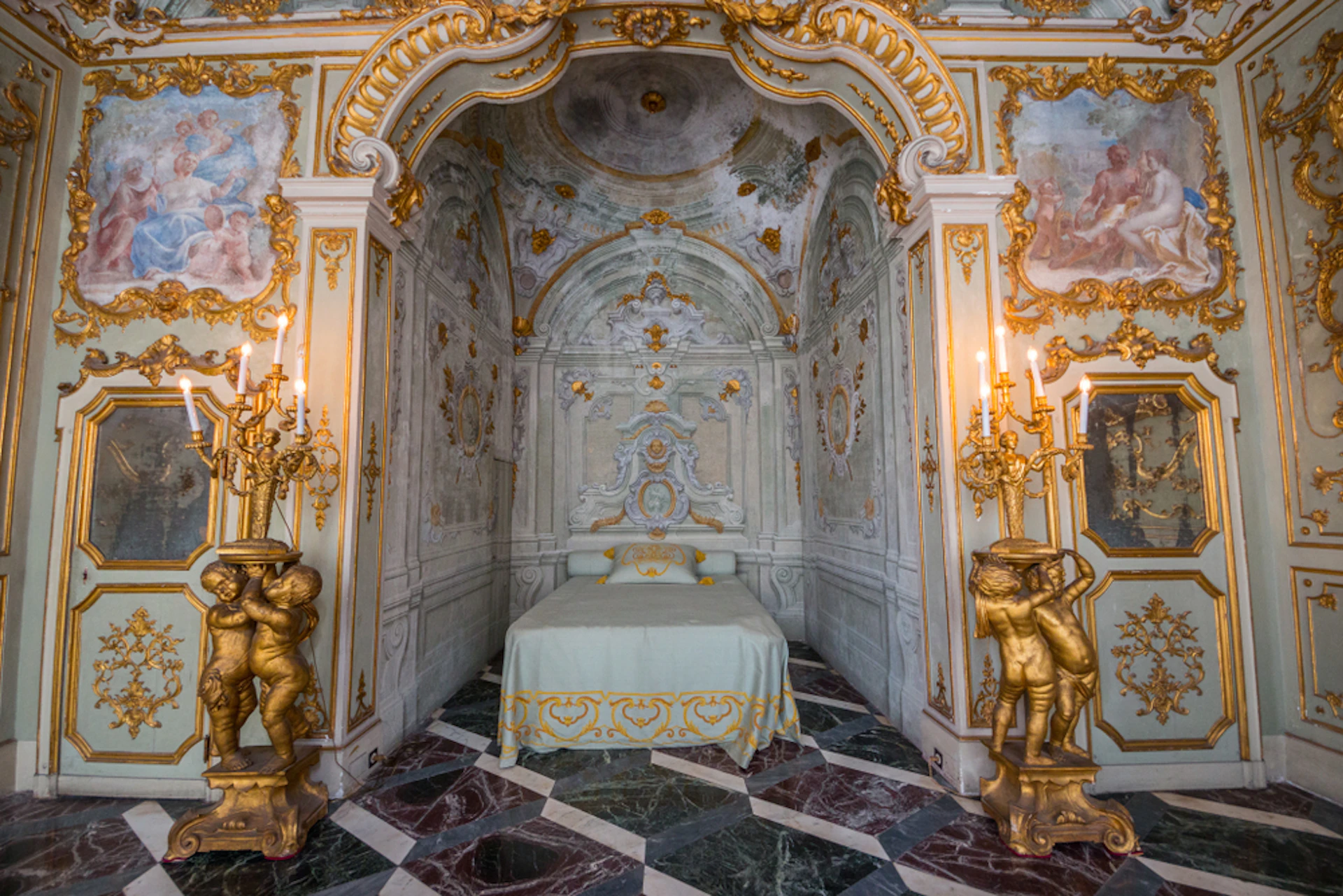 Luxurious Rococo-style room with ornate gold detailing, a canopied bed, marble floor, and golden candelabras held by cherub statues.