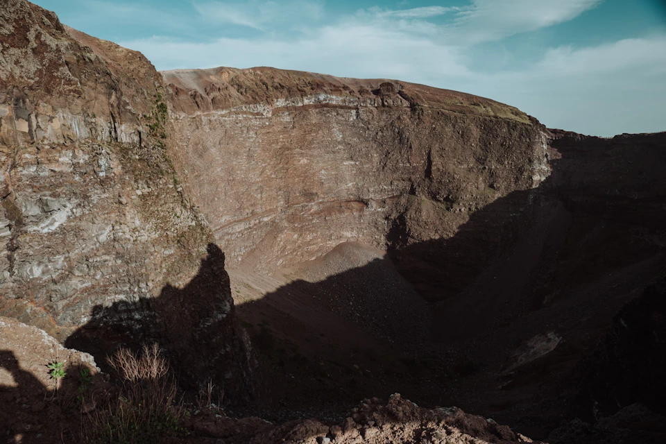 A rocky cliffside under a cloudy blue sky with stratified rock layers and a deep shadowed crater below.