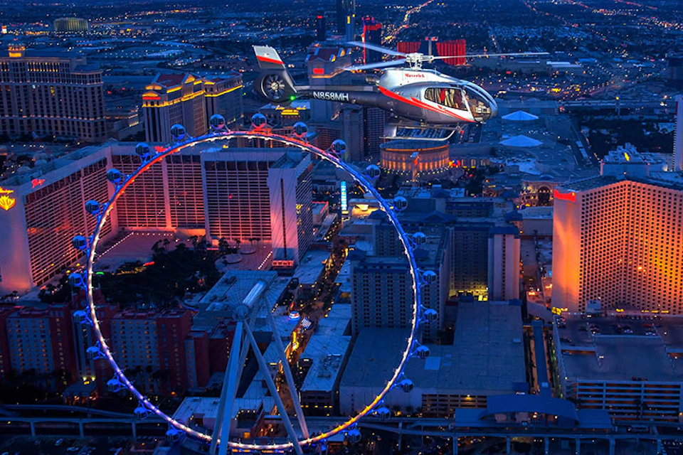 A helicopter flying near a large Ferris wheel at night over a city's brightly lit skyline.