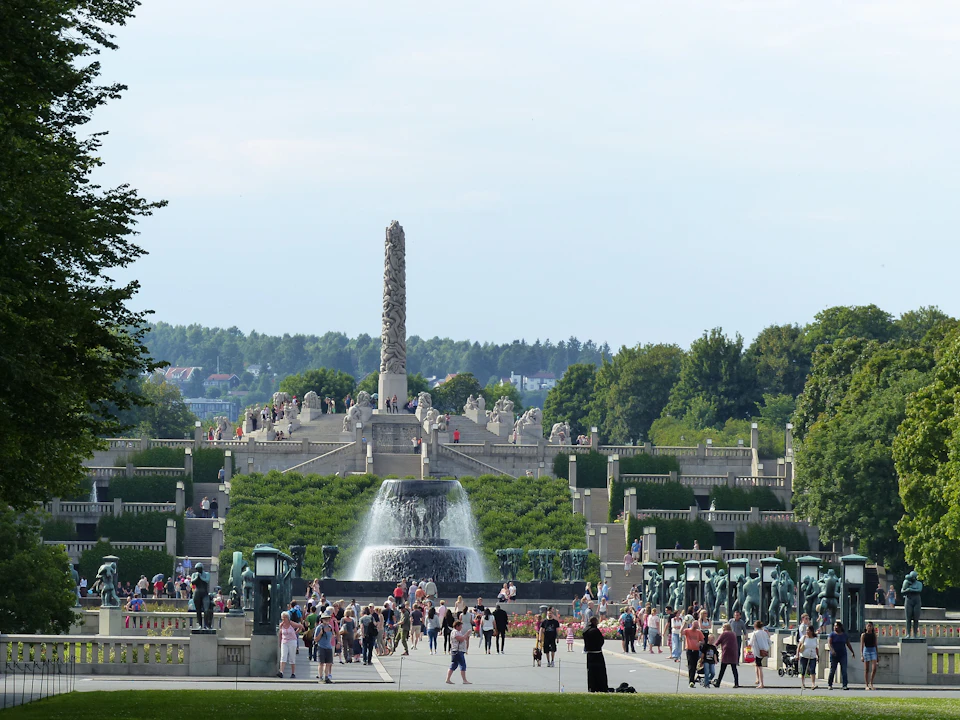A large outdoor park with a central monolith, a fountain in the foreground, and many people walking around. Trees and greenery surround the area.