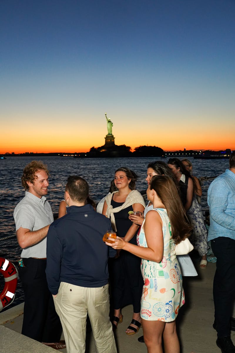 Μια ομάδα ανθρώπων που κοινωνικοποιούνται σε εξωτερικούς χώρους κοντά σε ένα σώμα νερού κατά το ηλιοβασίλεμα, με το Άγαλμα της Ελευθερίας ορατό στο παρασκήνιο.
