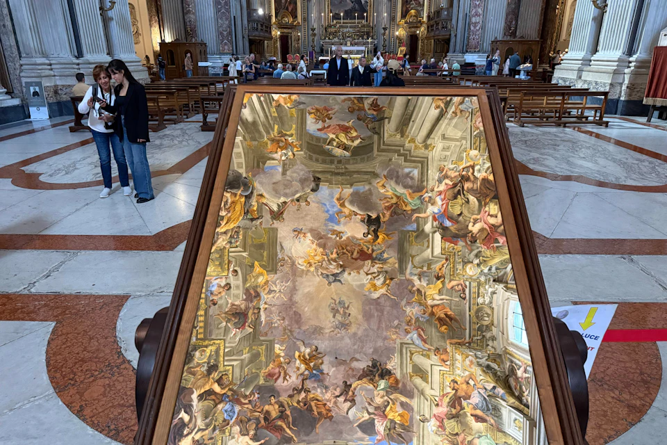 A reflective surface displaying the intricate ceiling fresco of a church, with visitors and pews in the background.