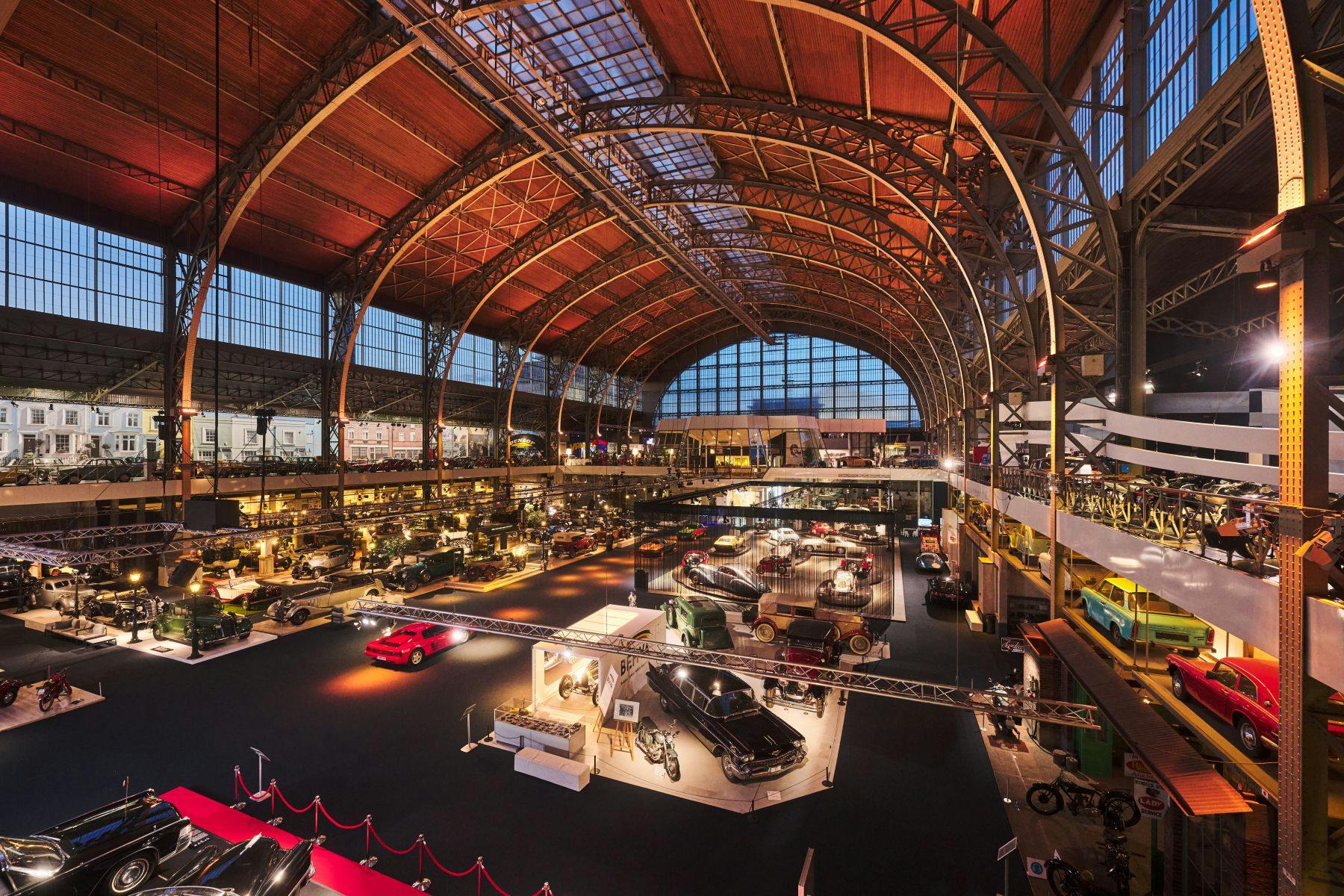 Un vaste hall de musée automobile avec de nombreux véhicules anciens, des vitrines illuminées et un plafond en bois voûté.