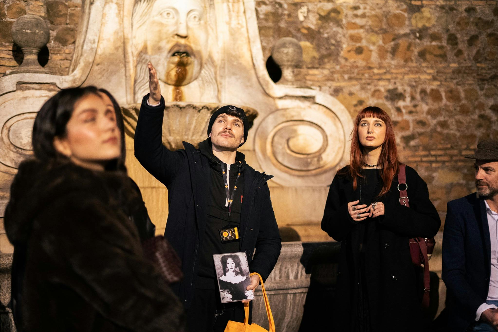A man and a woman stand near an ornate stone fountain, the man gesturing upwards while holding a photograph.