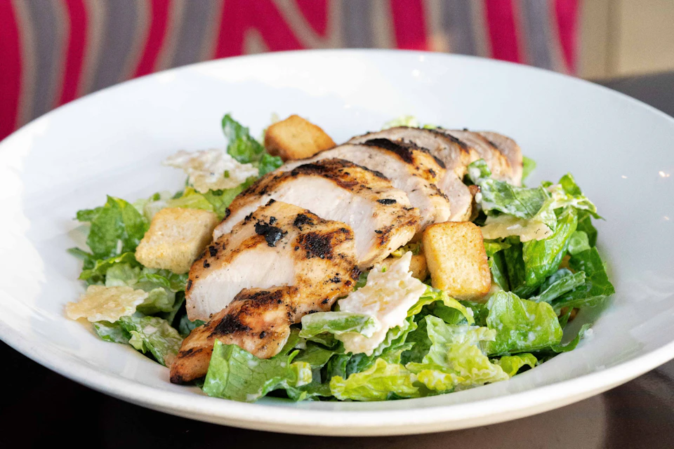 Grilled chicken breast on a bed of romaine lettuce, croutons, and Caesar dressing in a white bowl.