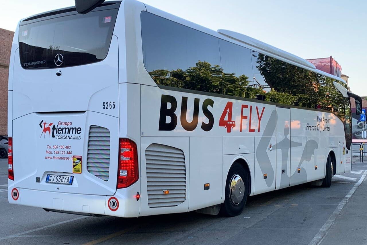 Weißer Bus mit "BUS 4 FLY"-Schild, der an einer Straße geparkt ist und in dessen Fenstern sich Bäume spiegeln.